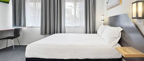 Une chambre d’hôtel avec un lit, un bureau et une chaise. Il y a des rideaux aux fenêtres et un tableau au mur.