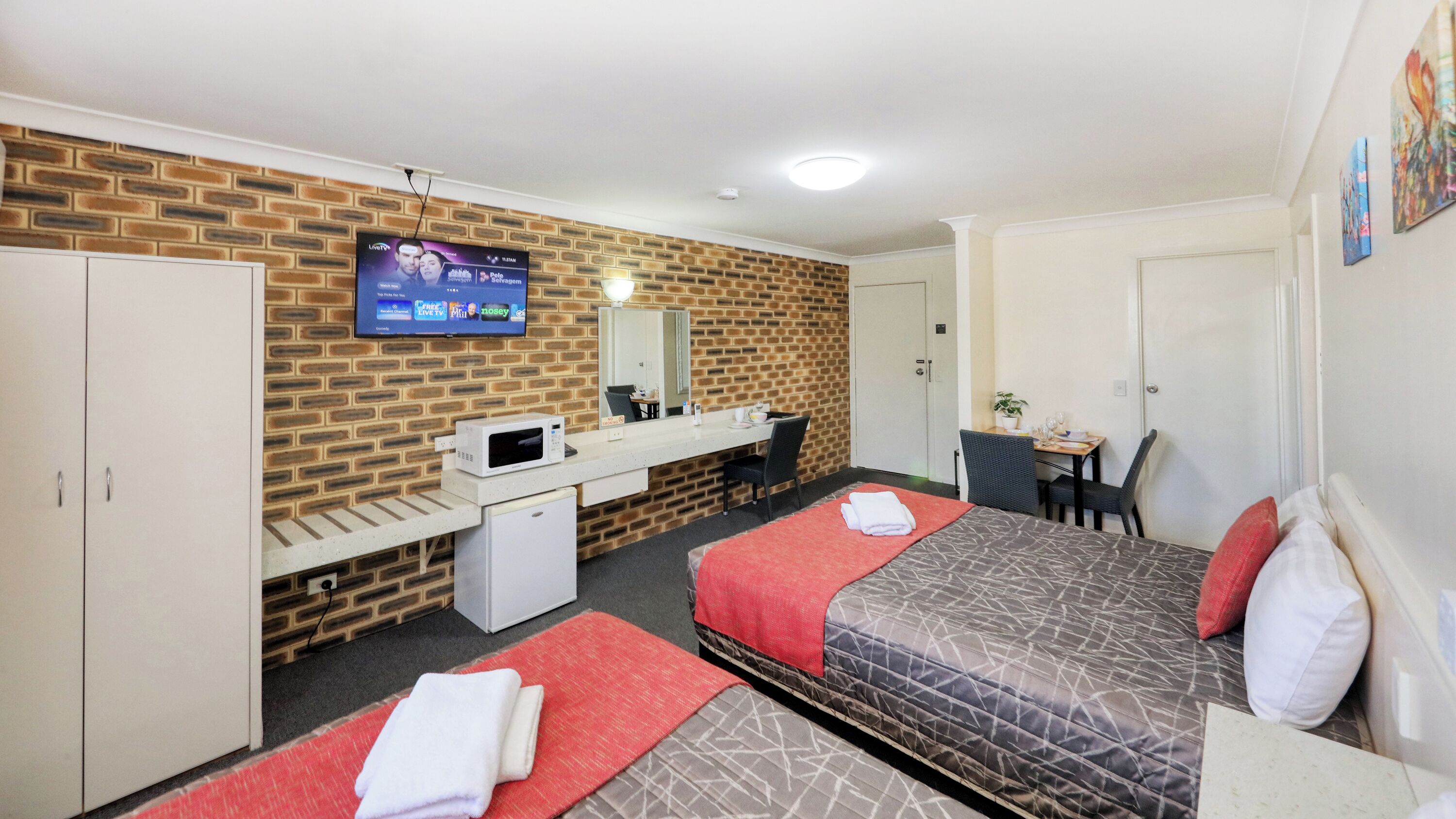 A hotel room with a brick-patterned wall, a flat-screen TV mounted above a desk, a microwave, a bed with a red and grey patterned bedspread, and a small table with a chair.