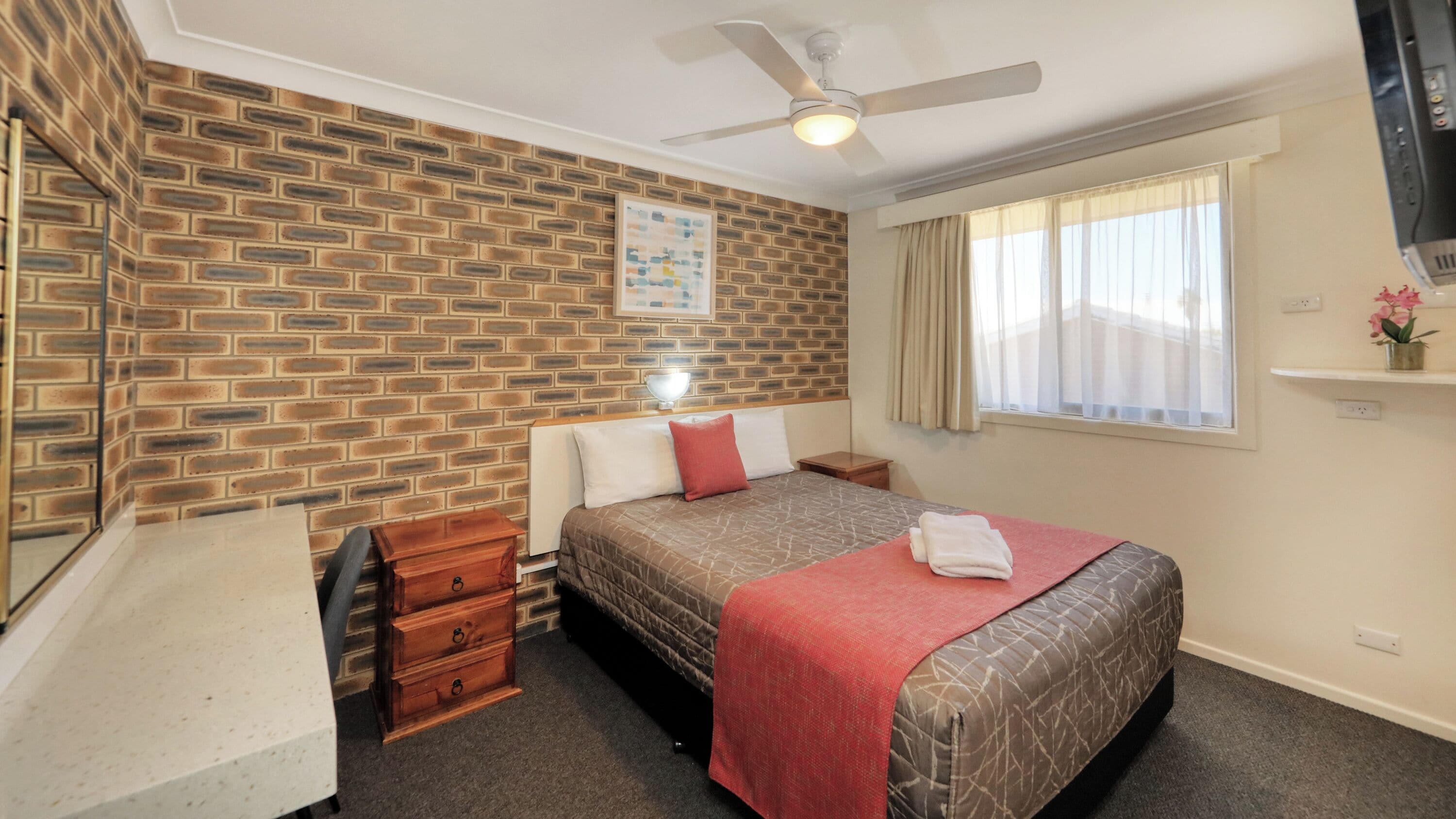 A bedroom with a brick-patterned wall, a bed with a red blanket, a ceiling fan, a bedside table, and a window with curtains.