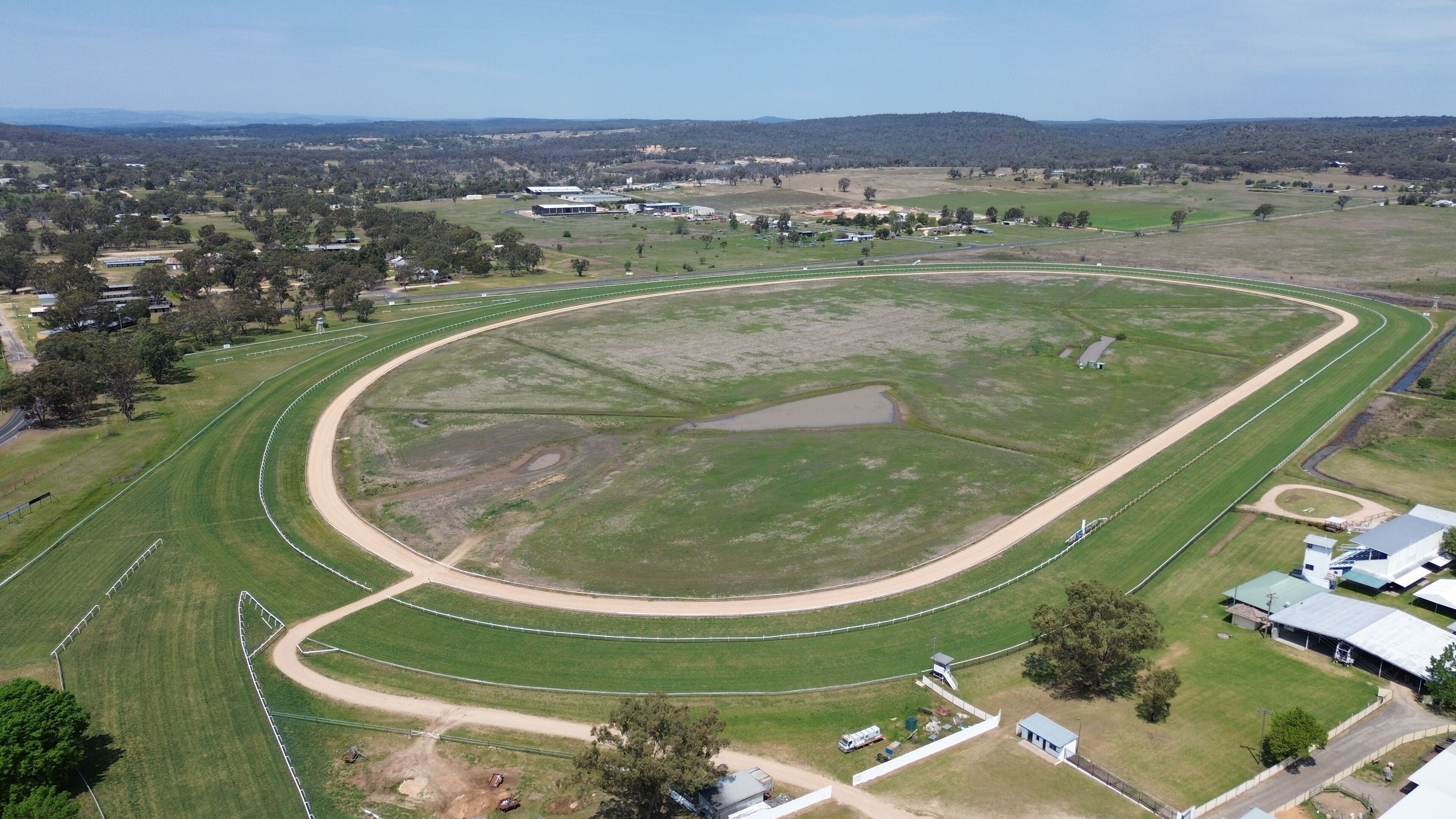 Eine Vogelperspektive auf ein großes, ovales Sportfeld mit einer Dirt-Track-Bahn, umgeben von Rasenflächen und vereinzelten Bäumen.