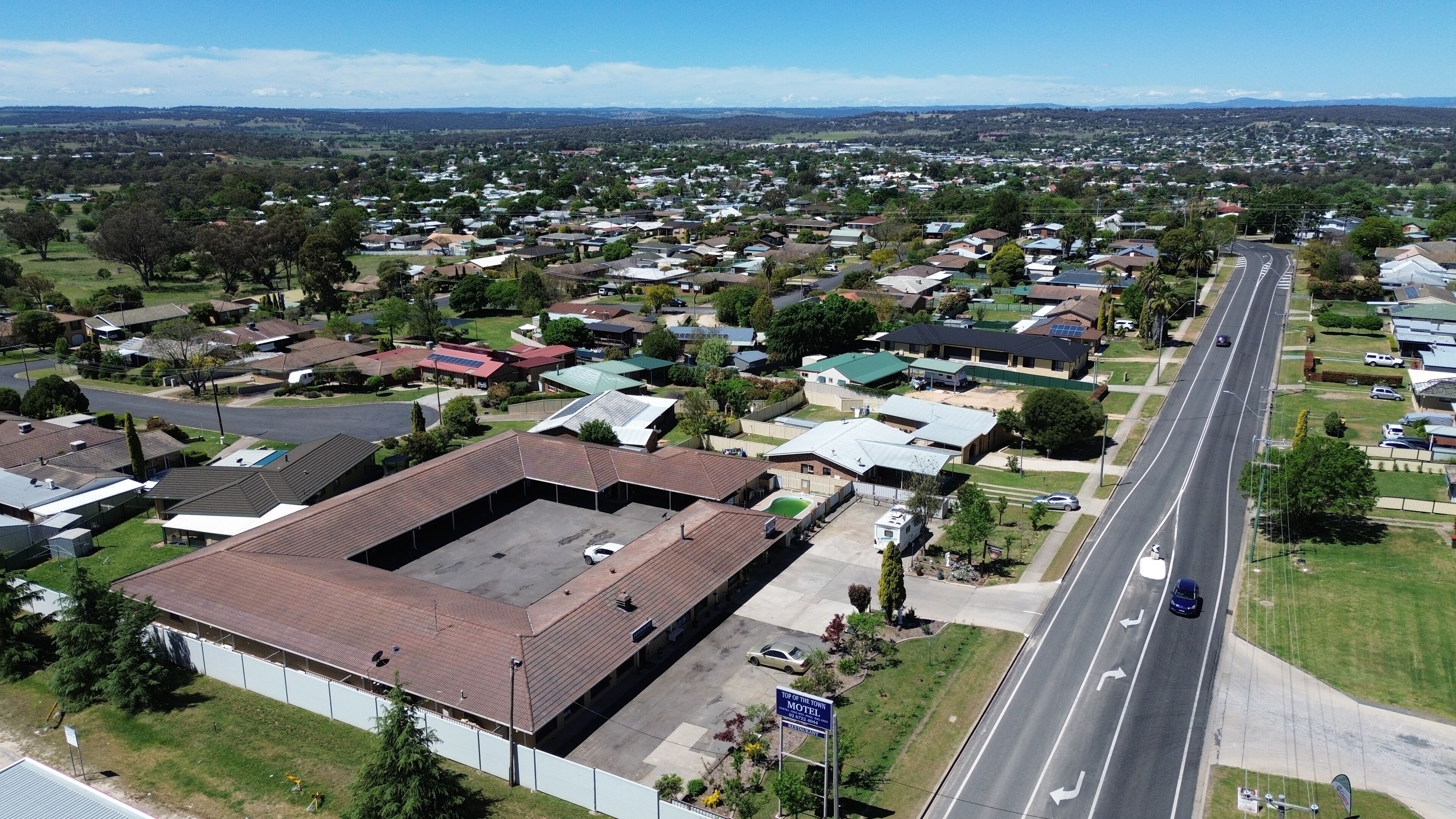 A motel with a large parking lot, a main building with a flat roof, and a road with a car passing by.