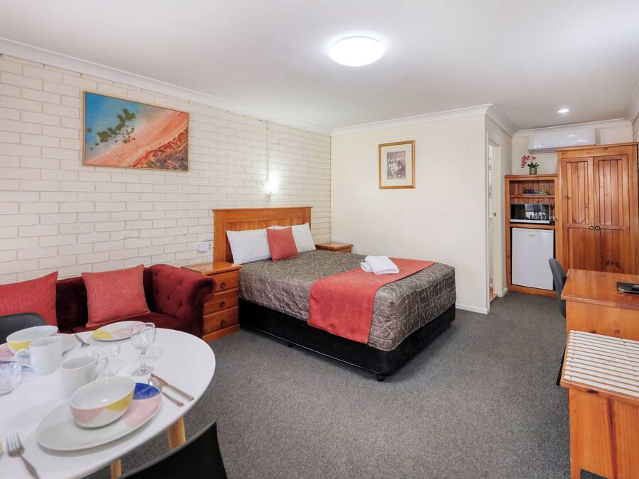 A compact hotel room with a bed, a small desk, a dining table with chairs, and a kitchenette.