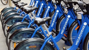 Una fila de bicicletas azules con la marca 'Melbourne', estacionadas en línea sobre una acera.