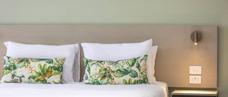 A neatly made bed with white linens and two pillows with a tropical leaf pattern. There is a bedside table with a light switch and a power outlet.