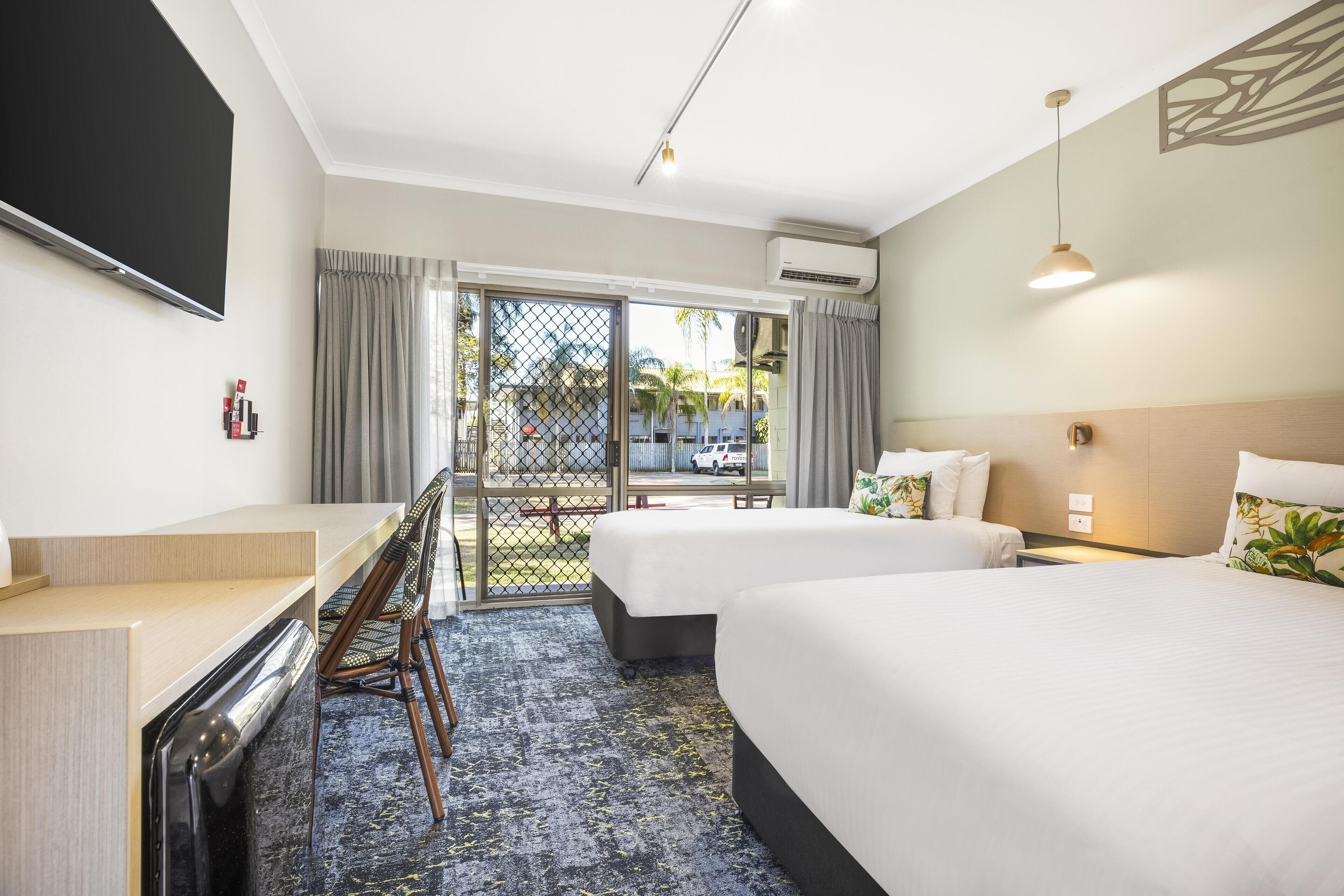 A hotel room with two beds, a desk, a TV, and a balcony with a view of palm trees.