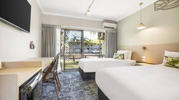 A hotel room with two beds, a desk, a TV, and a balcony with a view of palm trees.