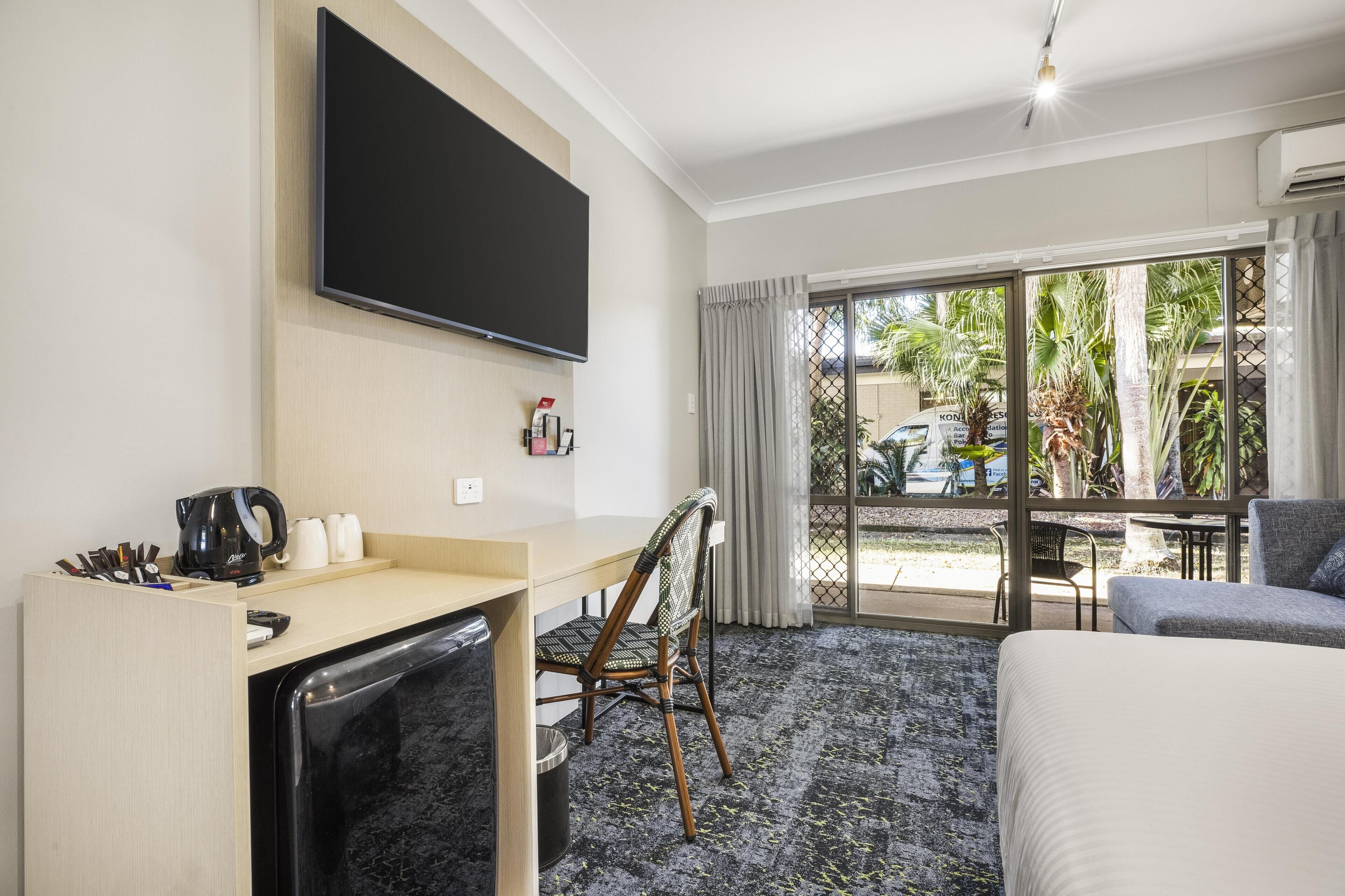 Habitación de hotel con cama, escritorio con cafetera, una silla, televisor montado en la pared y vistas al exterior a través de un amplio ventanal.