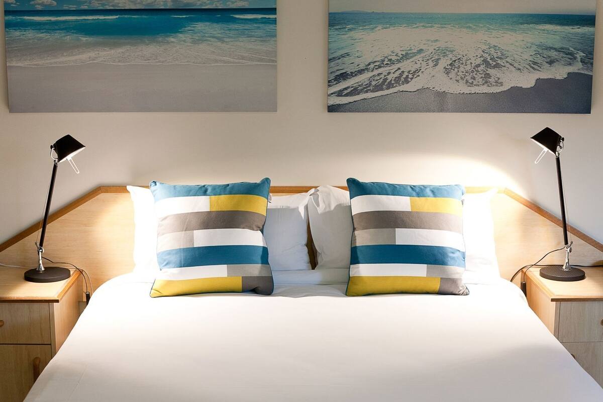 A bed with two pillows, two bedside lamps, and two wall-mounted pictures.