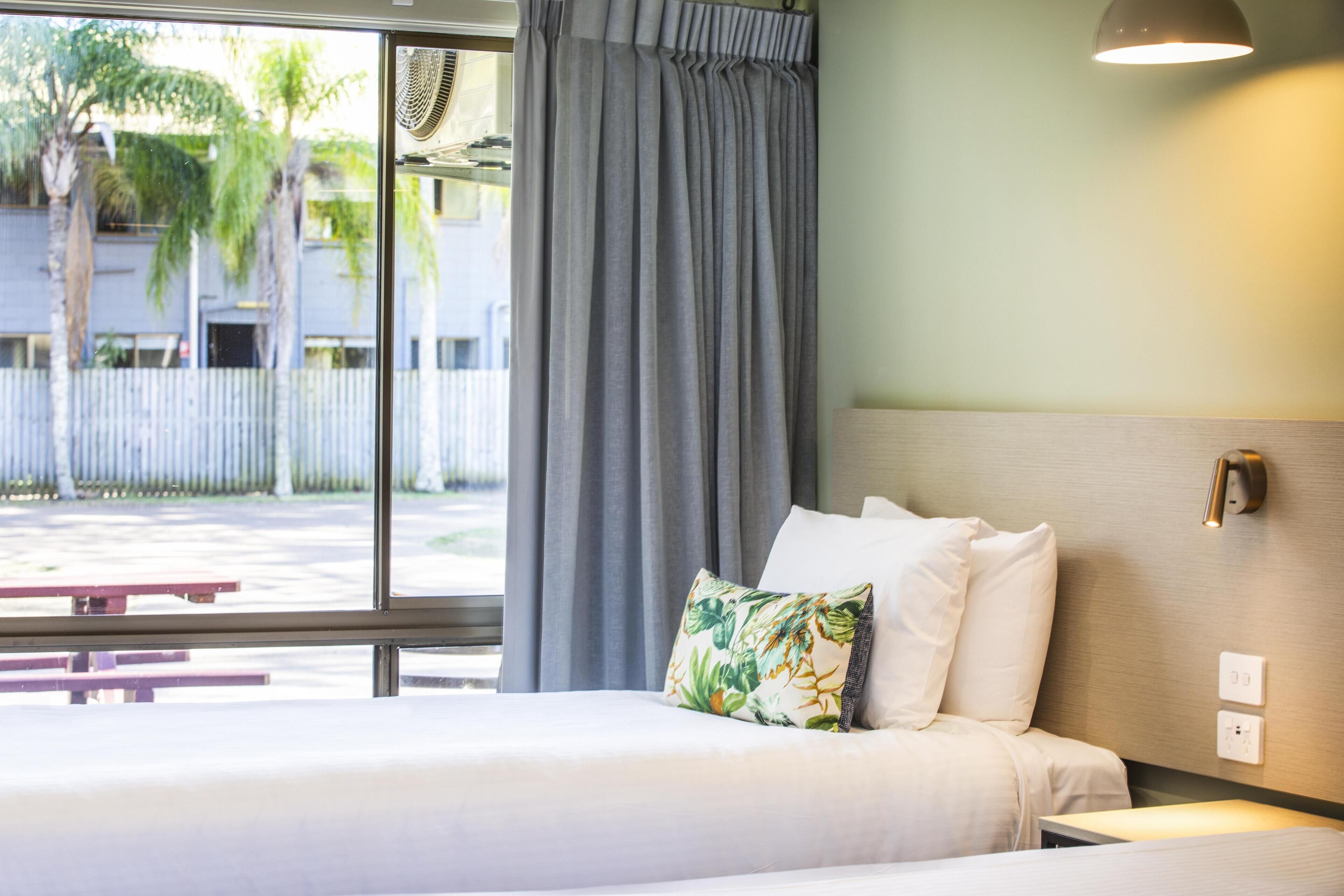A hotel room with a bed, a window with curtains, a bedside table, and a wall-mounted light.
