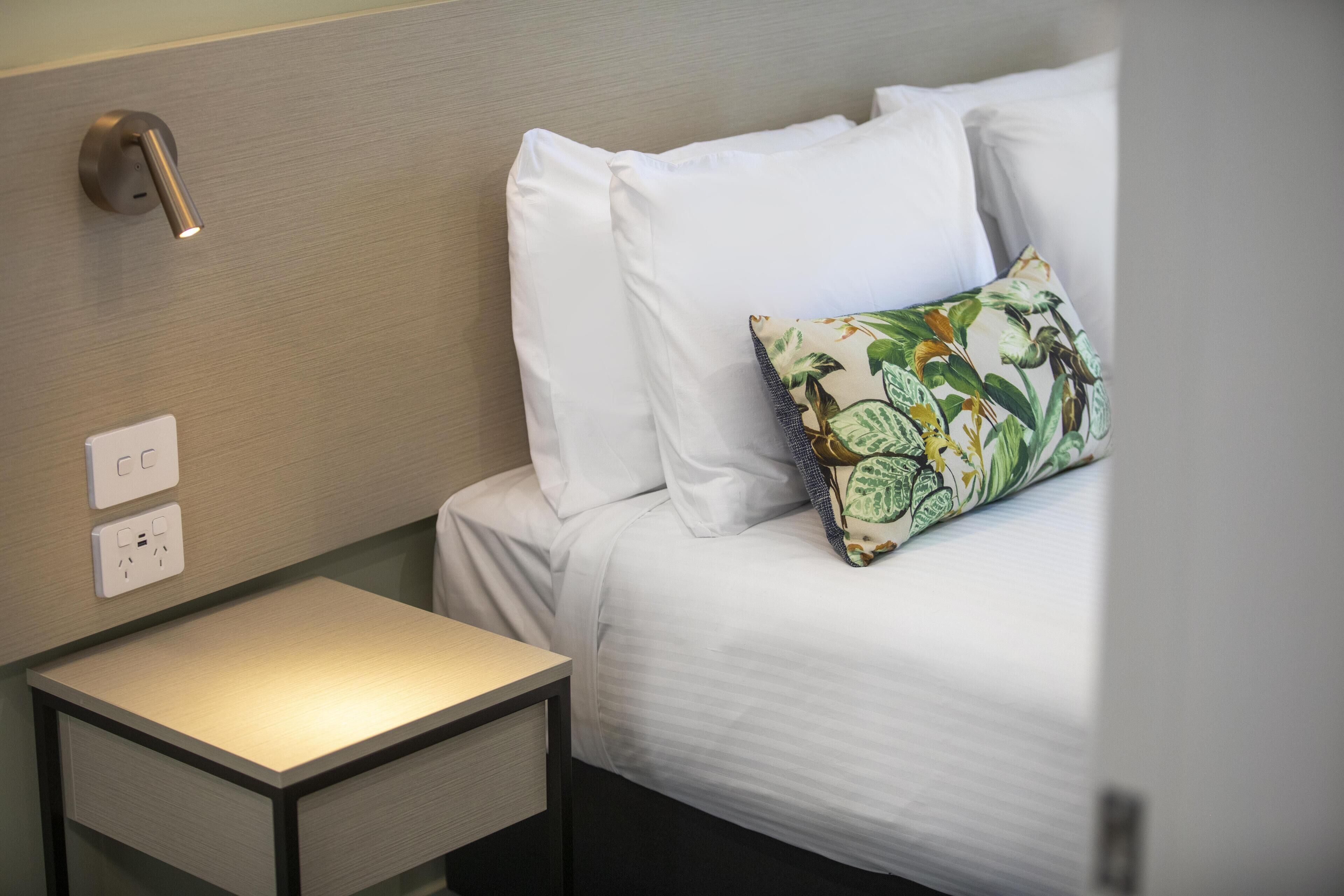 A neatly made bed with white linens and a floral pillow, a bedside table with a lamp, and a wall socket.