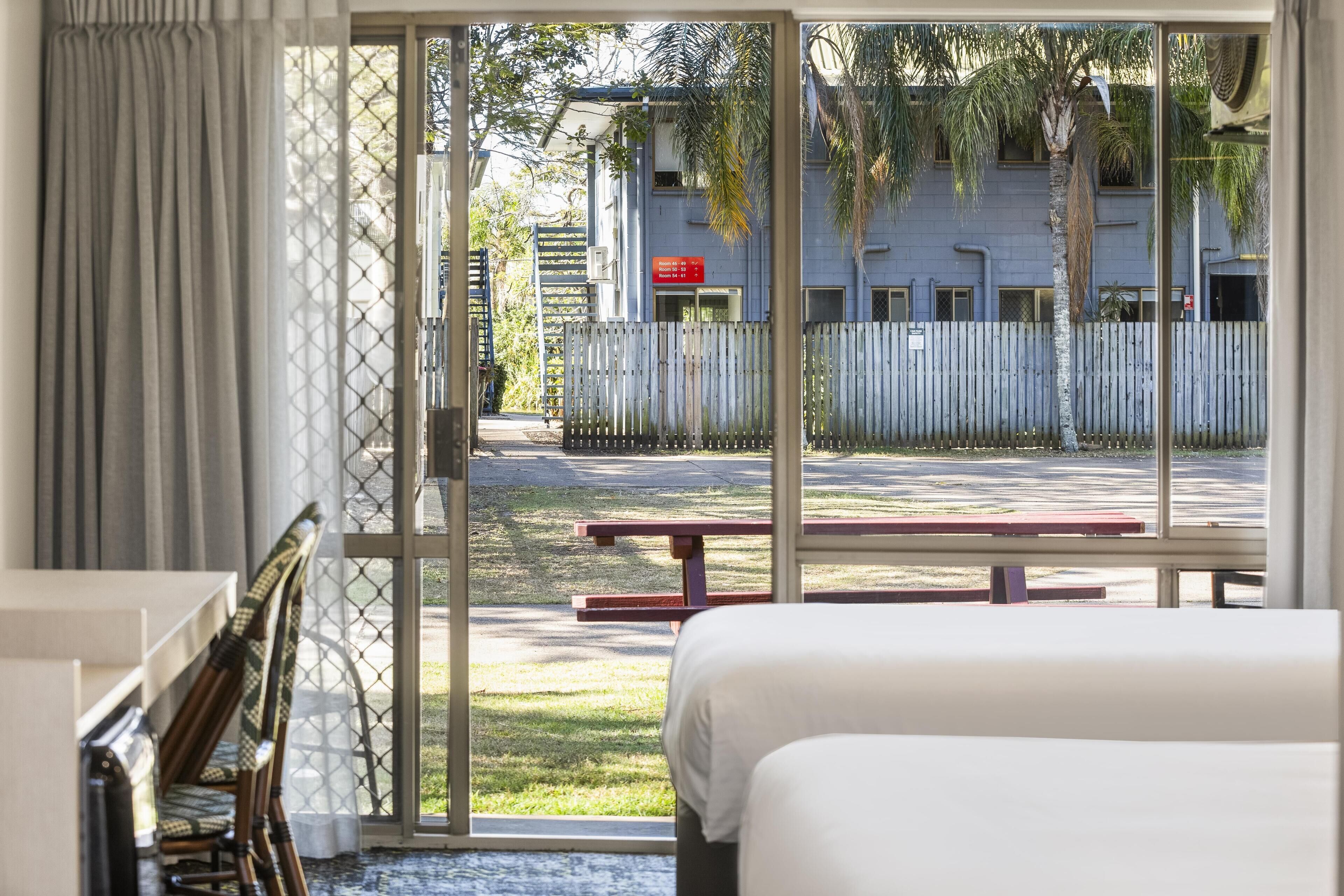 Habitación de hotel con cama, escritorio y silla. Se ve una ventana con vista a un edificio y una valla.