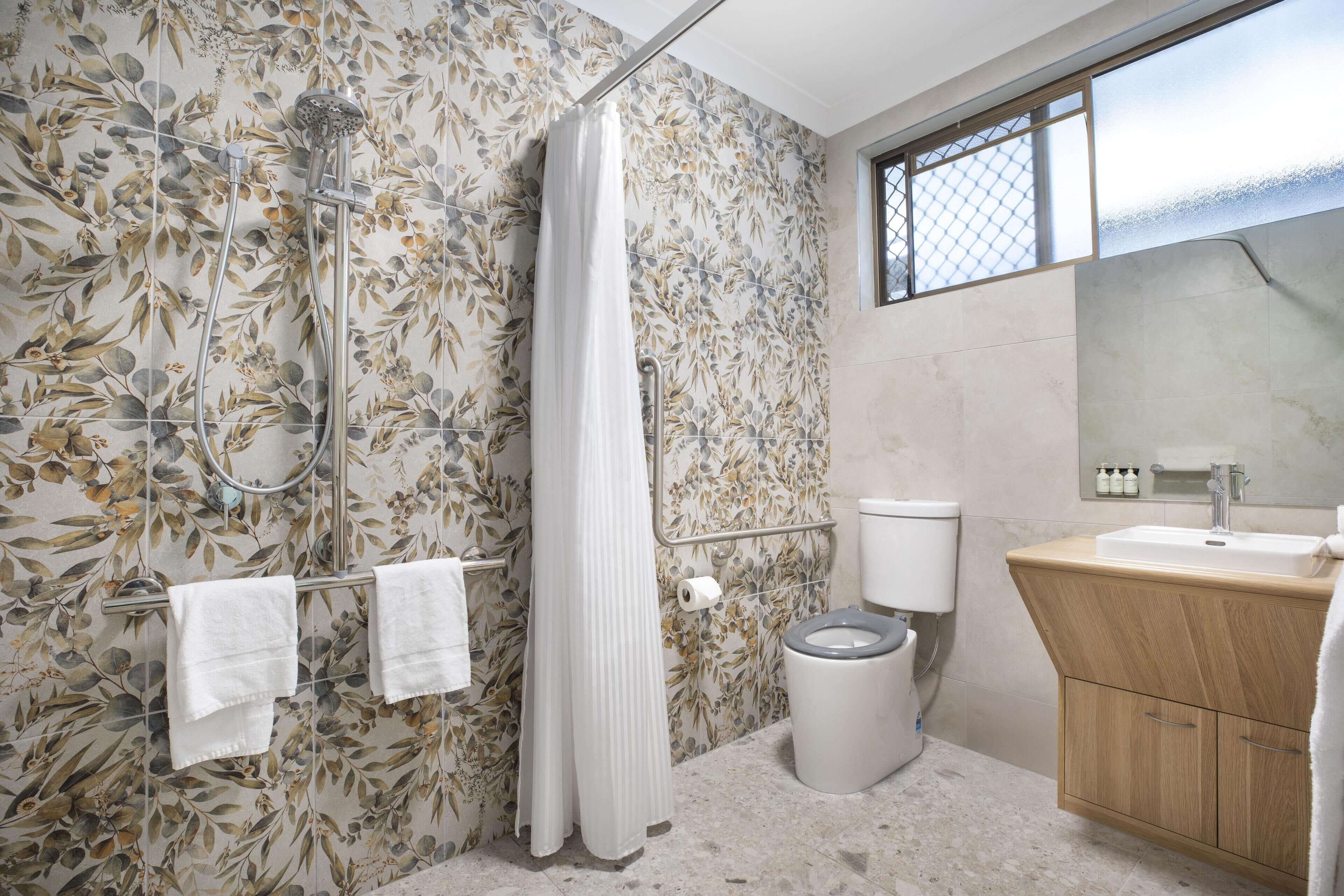 A bathroom with a shower, toilet, and sink, featuring floral wallpaper and a window.