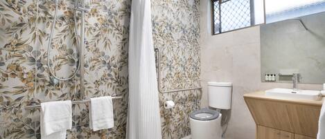 A bathroom with a shower, toilet, and sink, featuring floral wallpaper and a window.