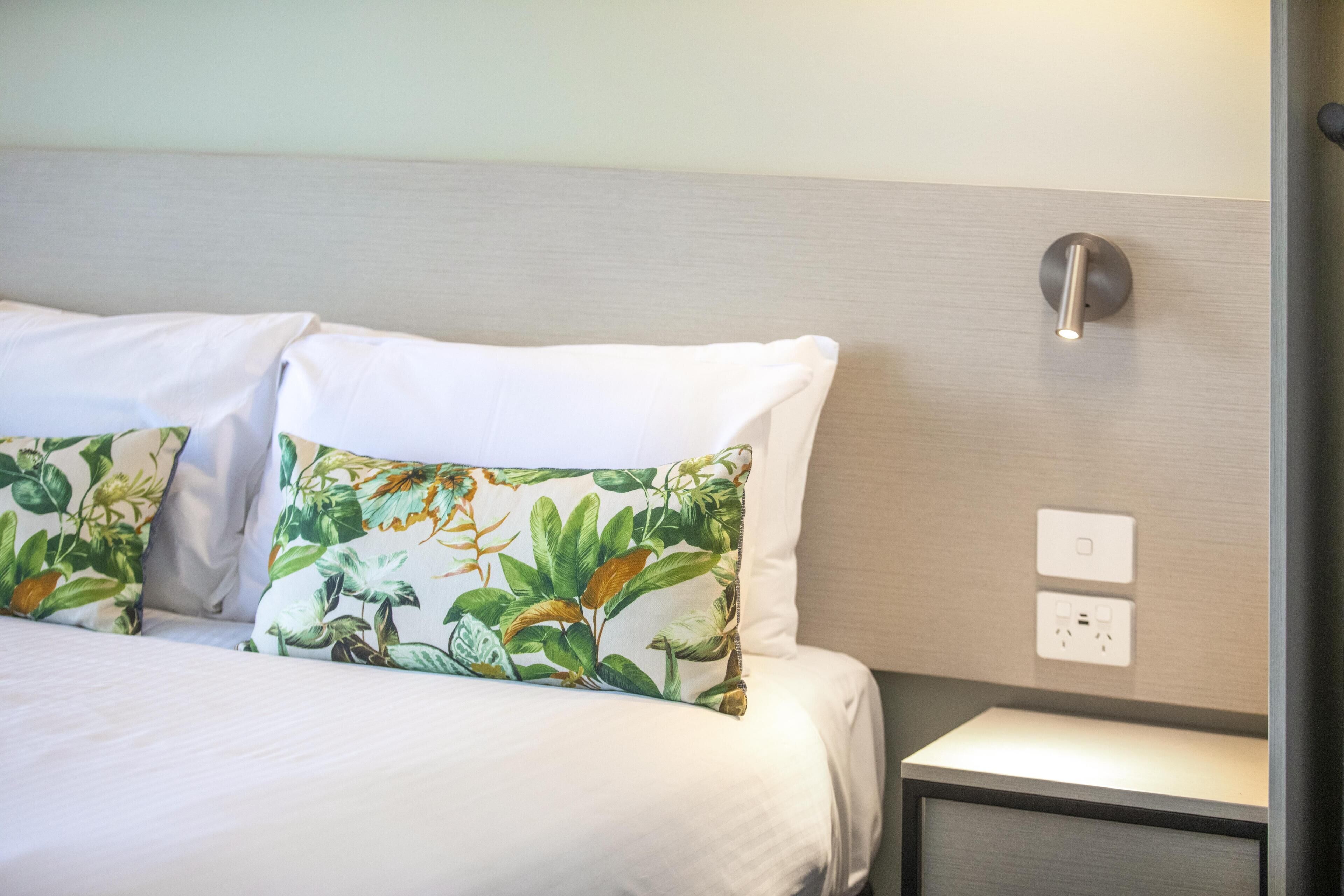 A neatly made bed with white linens and a floral pillow, a bedside table with a lamp, and a wall-mounted light switch.