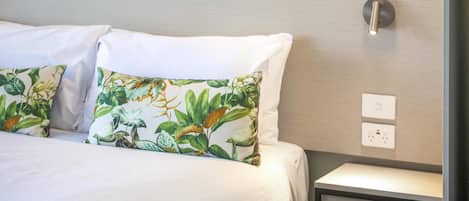 A neatly made bed with white linens and a floral pillow, a bedside table with a lamp, and a wall-mounted light switch.