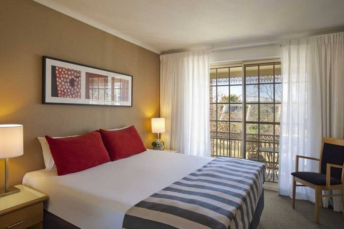 Ein Hotelzimmer mit einem großen Bett, zwei Nachttischlampen, einem gerahmten Bild an der Wand und einem Fenster mit Blick nach draußen.