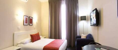 Habitación de hotel con cama, escritorio, silla, televisor y ventana con cortinas.