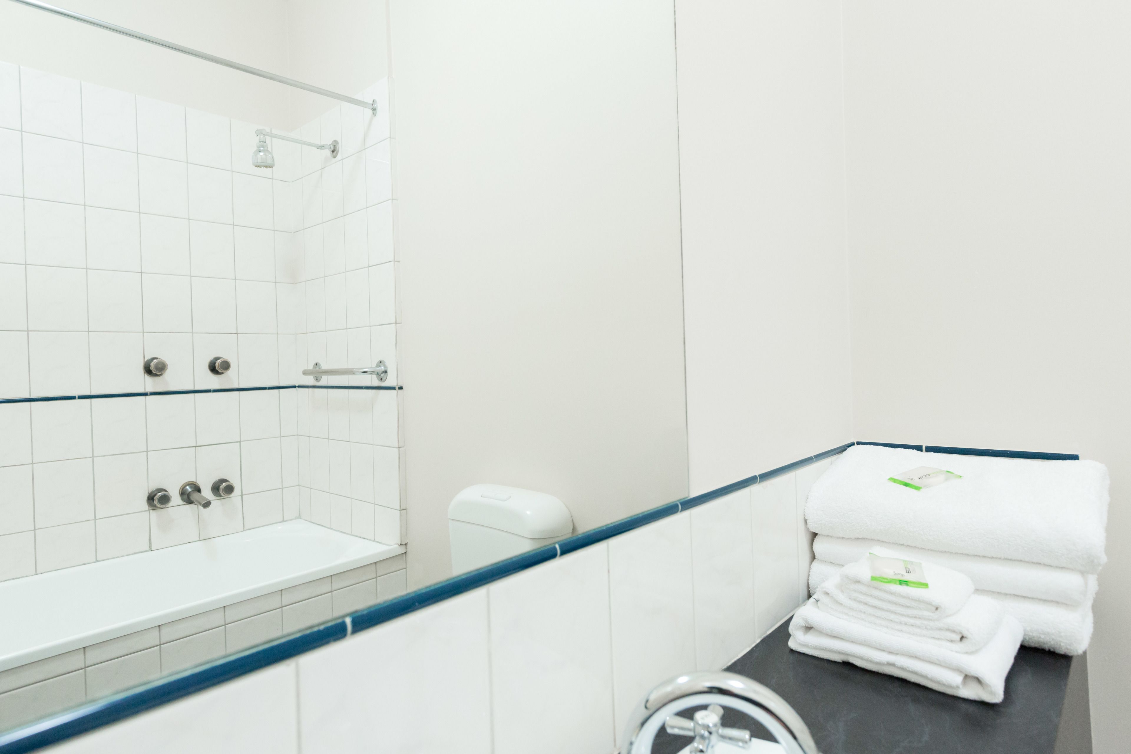 A bathroom with a glass shower enclosure, white tiles, a black countertop, and a stack of white towels.