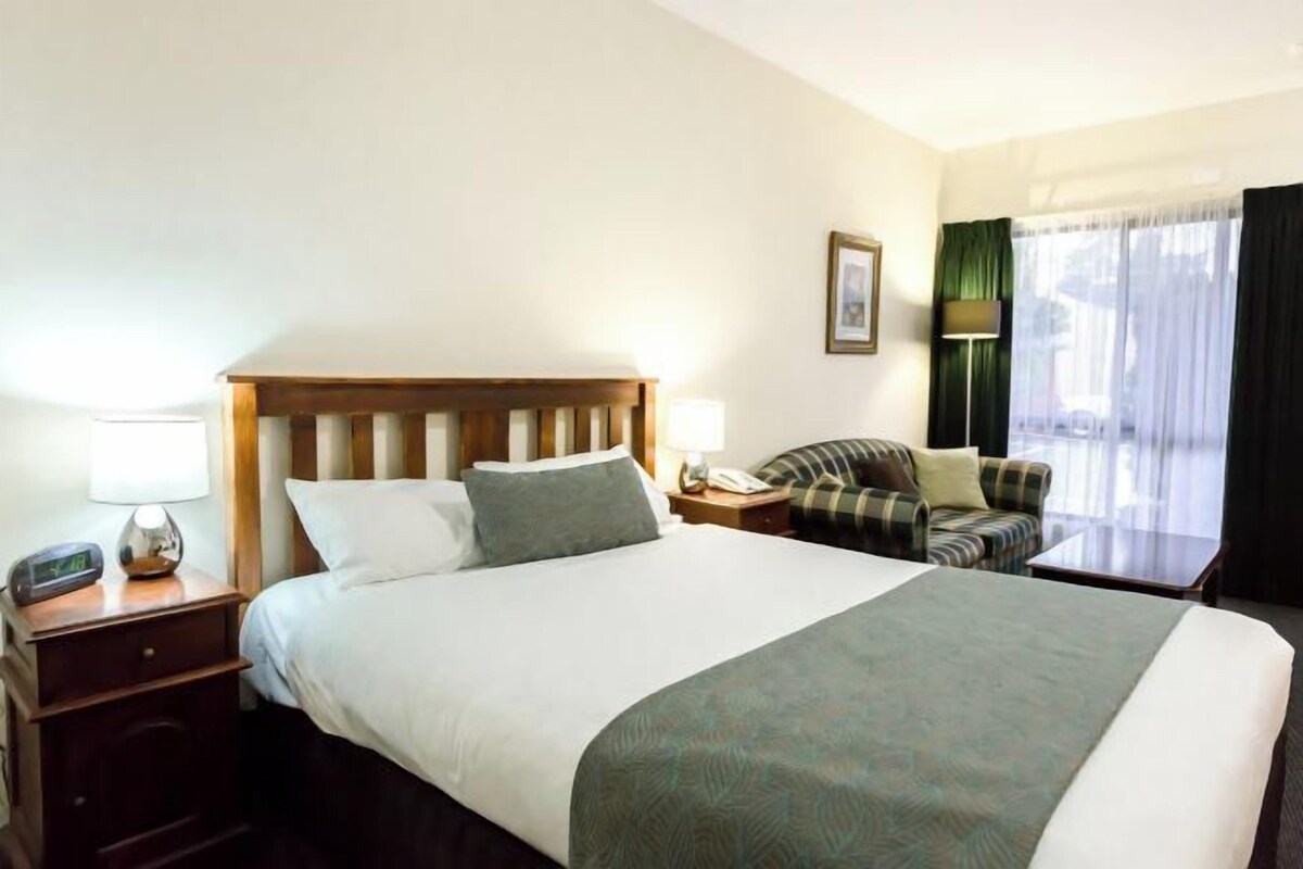 Ein Hotelzimmer mit einem großen Bett, einem Nachttisch mit Lampe, einem Sessel und einem Fenster mit Vorhängen.