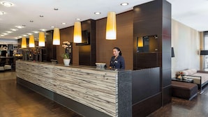 A modern reception desk with a wood-textured counter, a receptionist, and a seating area with a sofa and a small table. - NH Danube City (Vienna)