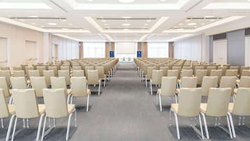 Un salón de conferencias con filas de sillas color beige, piso alfombrado y una pantalla grande al frente.