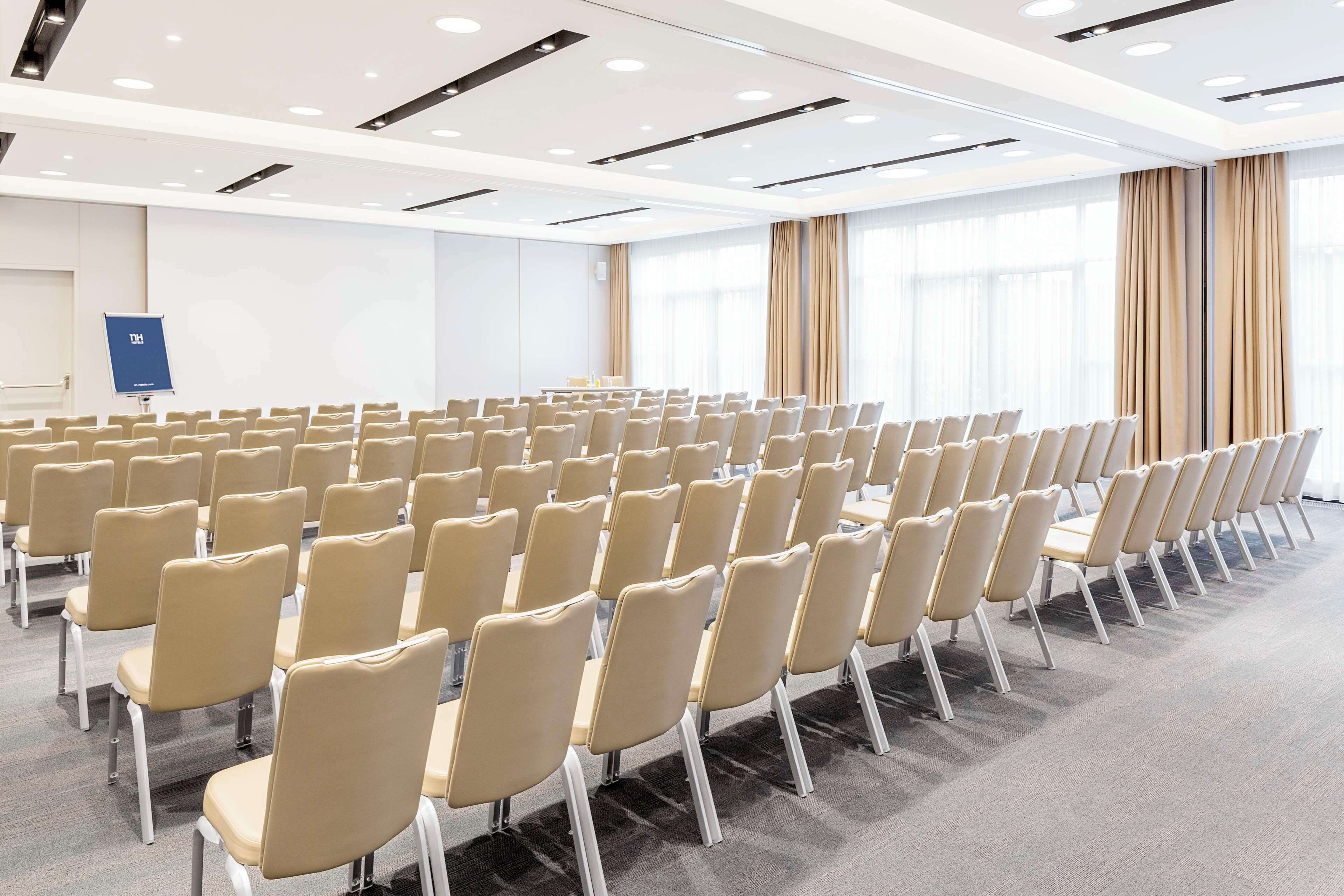 A conference room with rows of beige chairs, a presentation screen, and large windows with curtains.