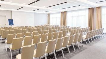 A conference room with rows of beige chairs, a presentation screen, and large windows with curtains.