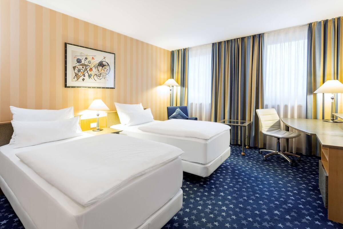 A hotel room with two beds, a desk, a chair, and a large window with curtains.