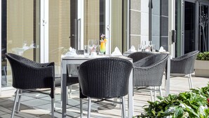 An outdoor dining area with wicker chairs and a table set for two, featuring a glass pitcher and wine glasses. - NH Danube City (Vienna)