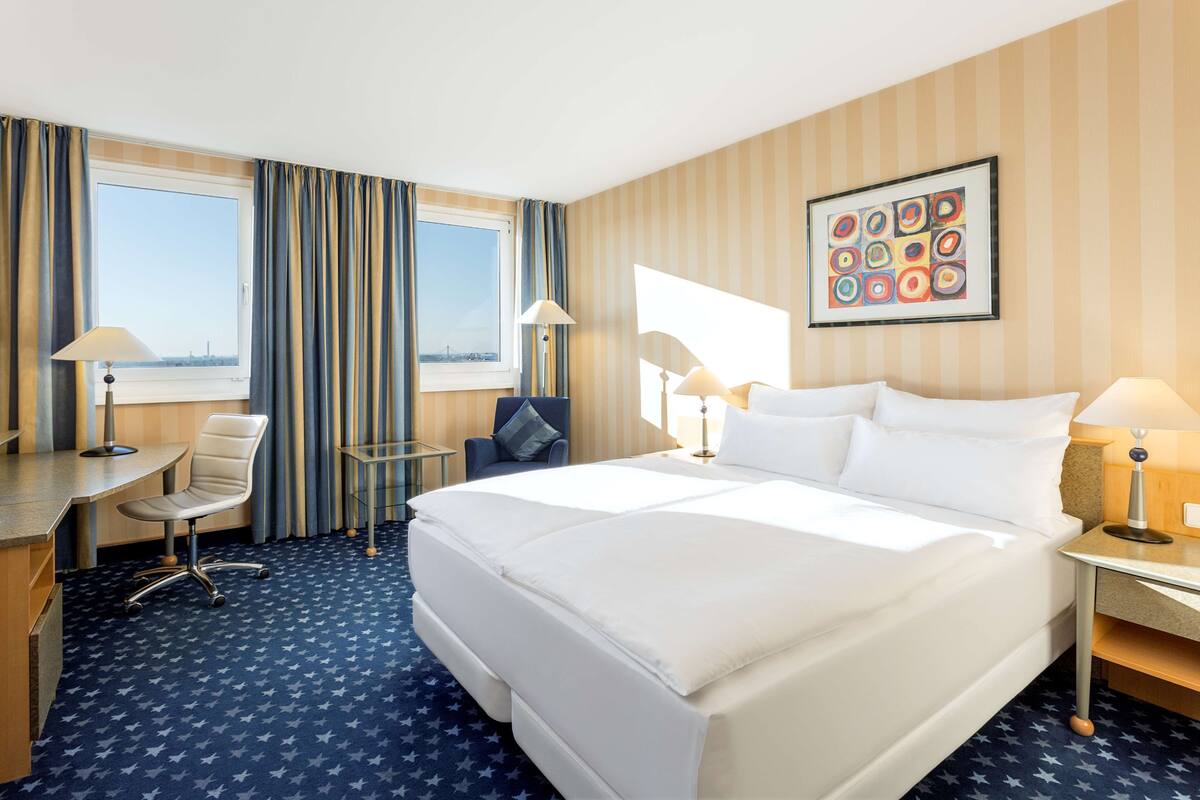 A hotel room with a large bed, two bedside tables, a desk, a chair, and a view of the city through the window.