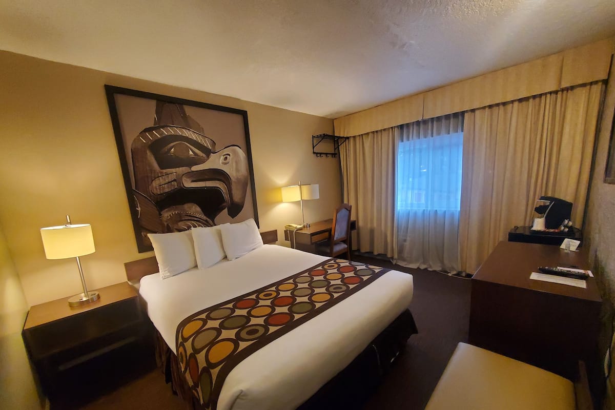 Una habitación de hotel con una cama grande, dos mesitas de noche, un escritorio, una silla, un cuadro abstracto de grandes dimensiones en la pared y una ventana con cortinas.