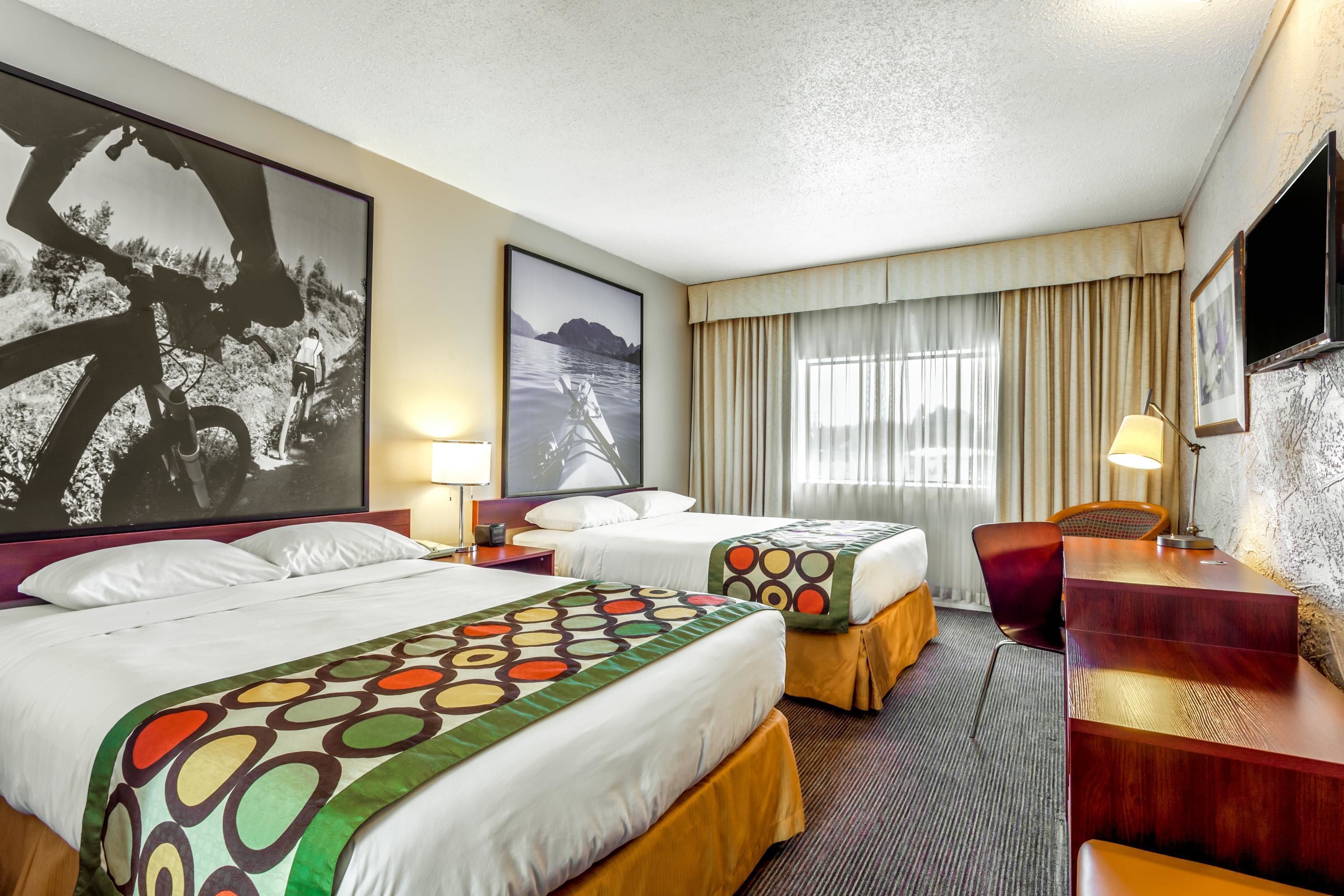 A hotel room with two beds, a desk, a chair, a TV, and large framed pictures on the walls.