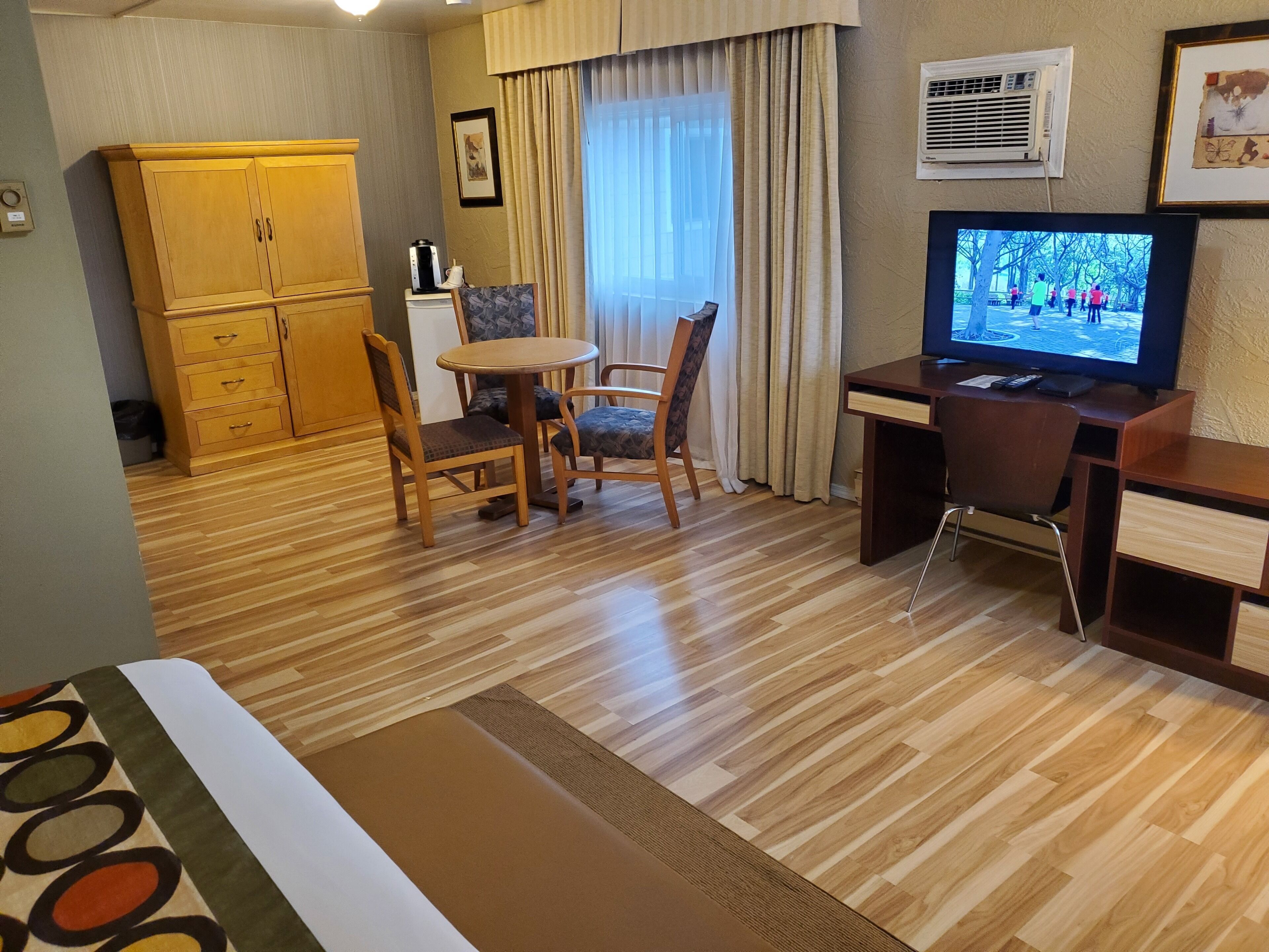 A hotel room with a wooden floor, a dining table with chairs, a TV, a wardrobe, and a window with curtains.