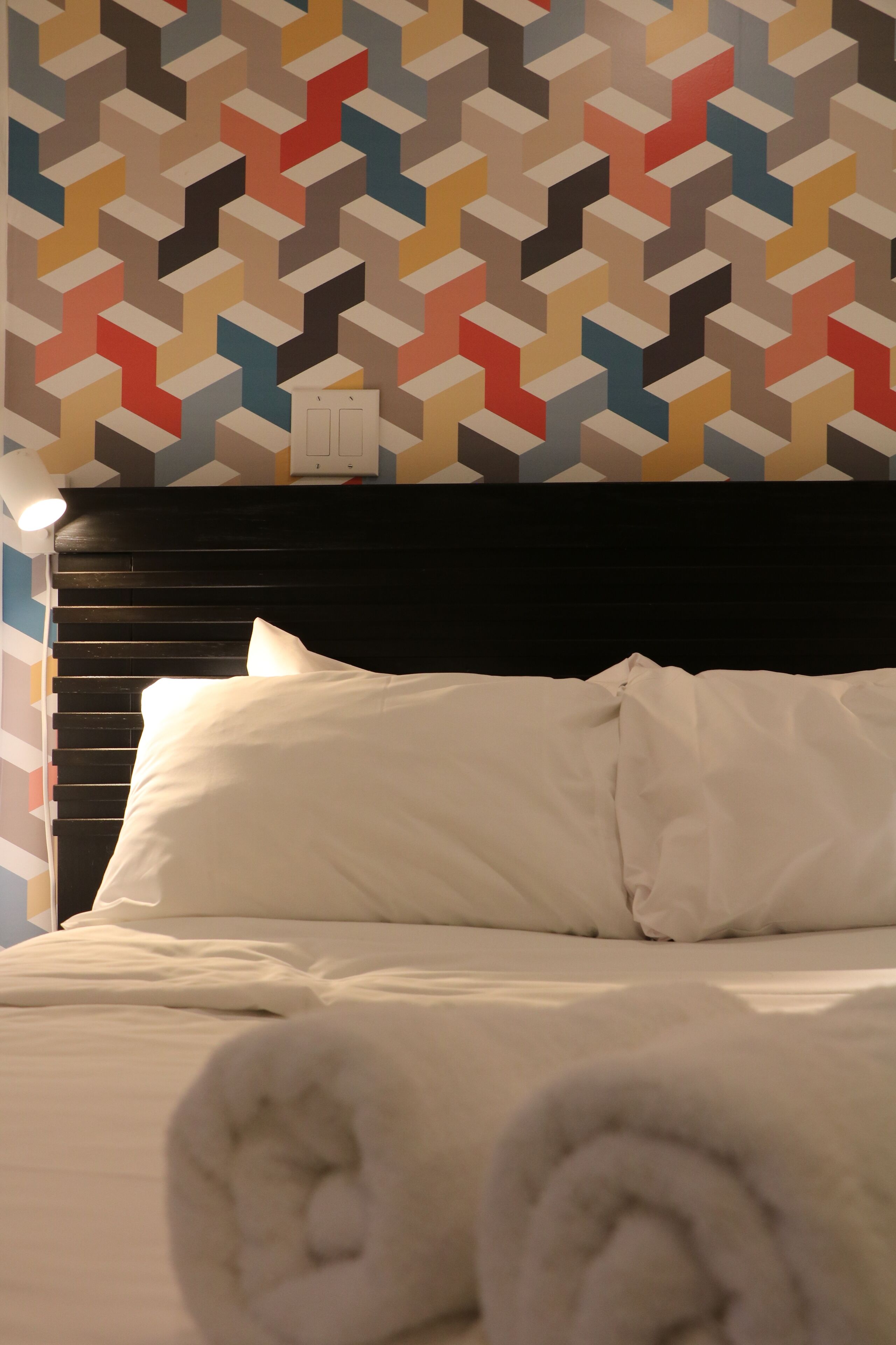 A neatly made bed with white linens and pillows, a headboard, and a patterned wall behind it.
