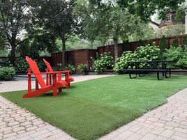 Een verzorgde tuin met kunstgras, een picknicktafel en rode Adirondack-stoelen.
