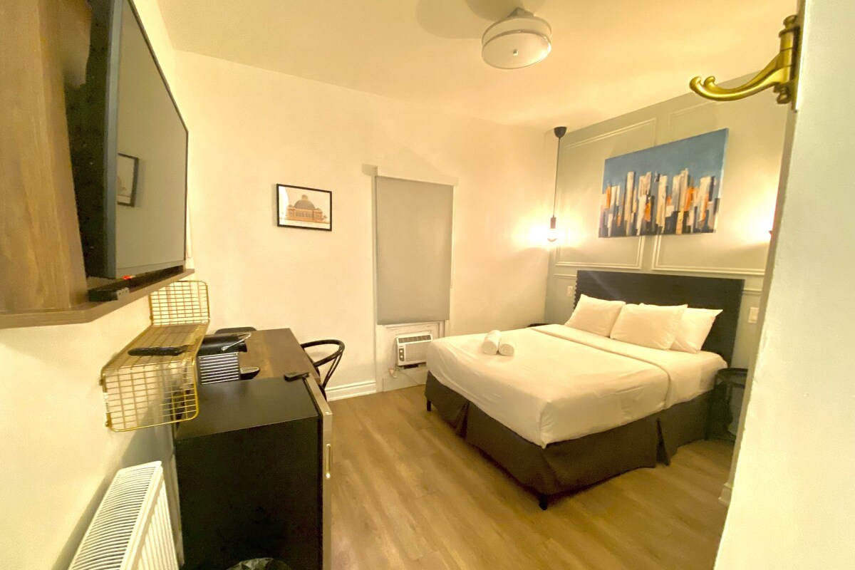 Habitación de hotel con cama, escritorio con computadora, televisión, radiador y un cuadro en la pared.