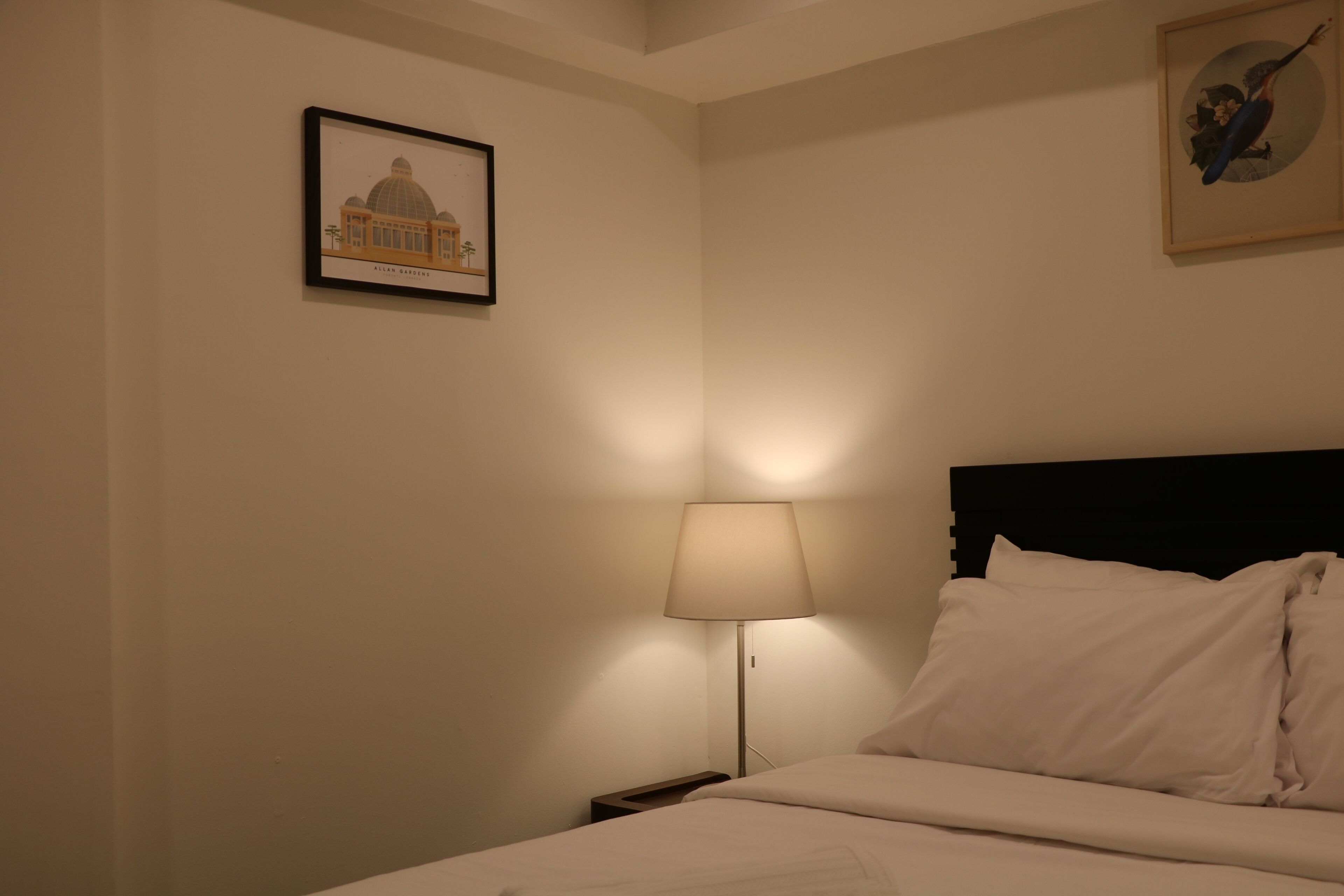 Hotellihuone, jossa on sänky, yöpöytä, lamppu ja kaksi kehystettyä kuvaa seinillä.