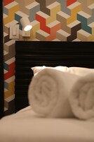 Een net opgemaakt bed met opgerolde witte handdoeken, een muur met een geometrisch patroon, en een wandlamp.