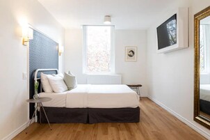 Een slaapkamer met een bed, een raam, een radiator, een tafeltje en een aan de muur hangend schilderij.