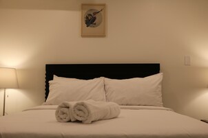 Een netjes opgemaakt bed met witte lakens en twee opgerolde handdoeken, een zwart hoofdbord, en een ingelijst schilderij aan de muur.