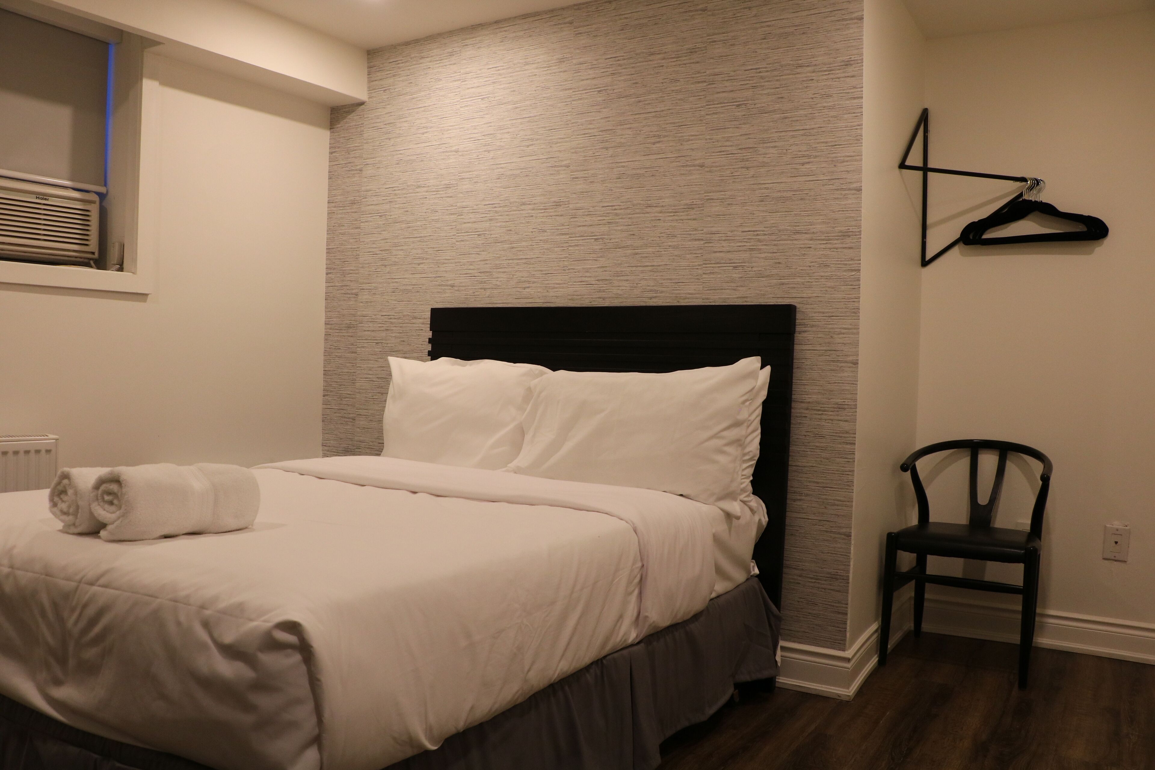 Una cama bien tendida con sábanas blancas y una toalla enrollada, un cabecero tapizado en negro, una silla de madera y un perchero de pared.