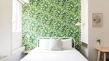 A bedroom with a green leafy wallpaper, a white bed, and a small bedside table with a lamp.