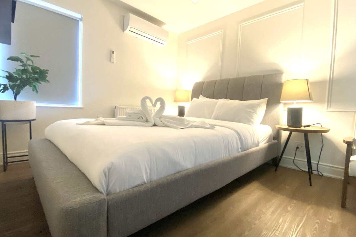 Una cama bien tendida con una toalla con forma de cisne, una lámpara de noche y una planta en maceta.