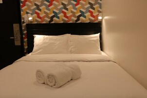 Een net opgemaakt bed met witte lakens en een opgerolde handdoek, een zwart hoofdeinde en een wand met een kleurrijk geometrisch patroon.