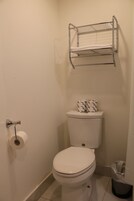 Een kleine badkamer met een toilet, een toiletrol, een metalen plank boven het toilet en een vuilnisbak.