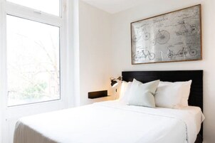 Een slaapkamer met een groot raam, een bed met witte beddengoed en een fiets-thema kunstwerk aan de muur.