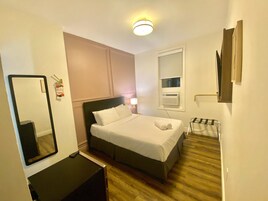 Habitación de hotel con cama, mesita de noche, espejo y un pequeño rincón de descanso con una silla.