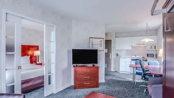 Habitación de hotel con cama, televisor, cómoda de madera, escritorio con silla, kitchenette y baño visible a través de una puerta abierta.