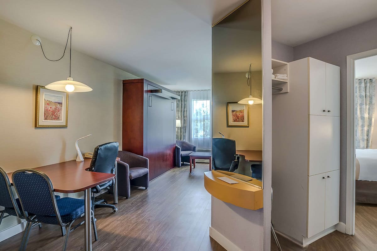 A modern hotel room with a dining area, a sofa, a desk, and a wardrobe.