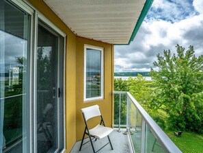 A balcony with a white chair, a glass door, and a view of greenery and a lake.